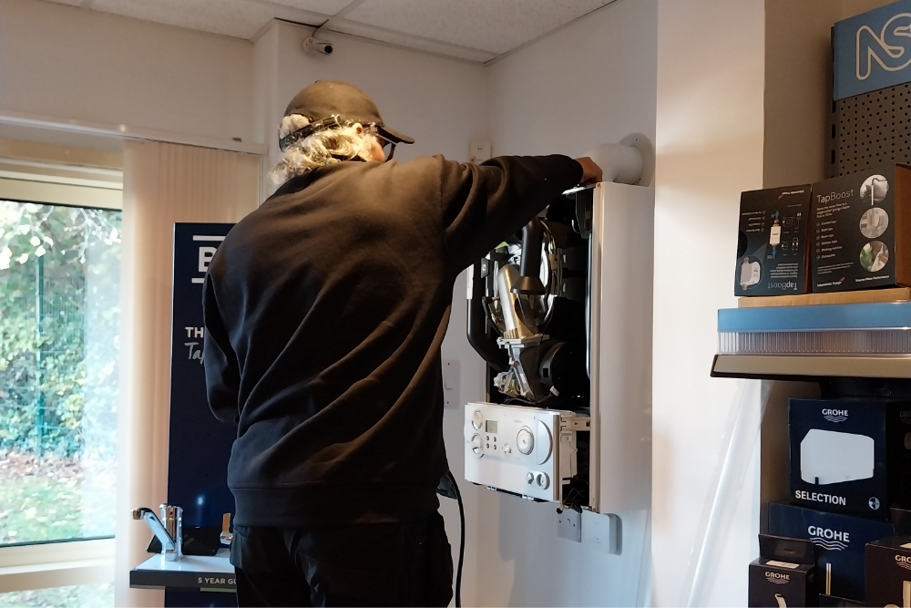 Servicing in progress: A technician works on the internal components of a wall-mounted boiler in a showroom filled with heating system displays.