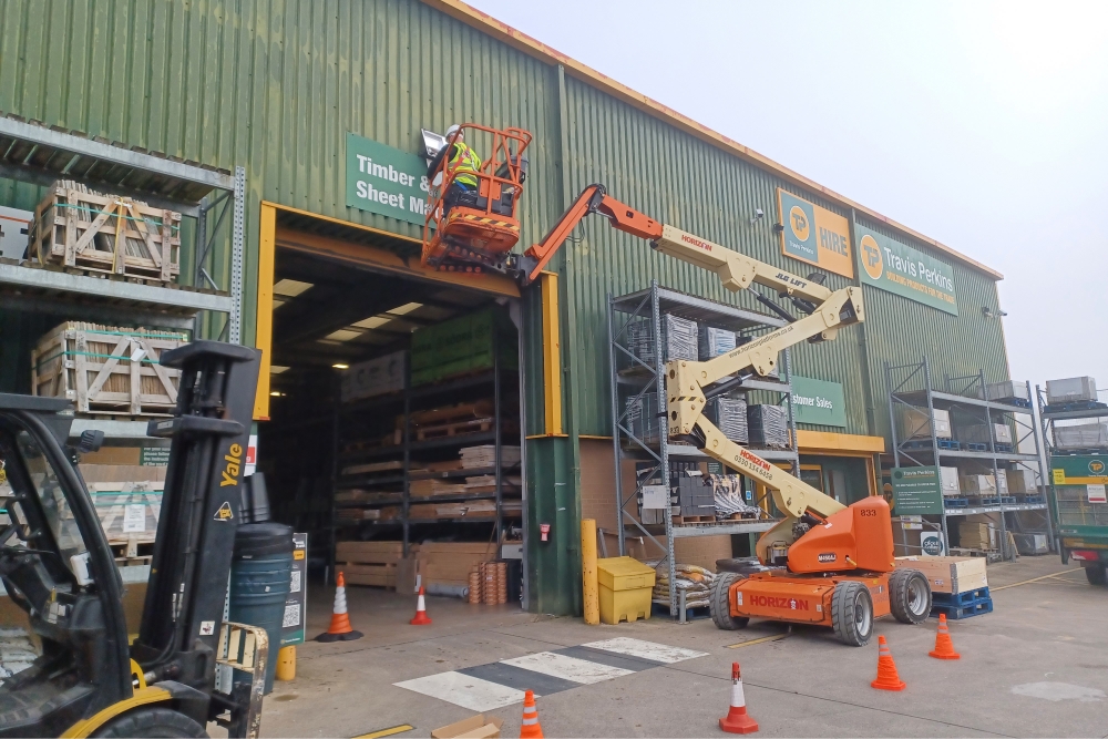 Exterior maintenance in progress at a Travis Perkins timber and sheet material warehouse, with a boom lift in use and safety measures in place around stored construction supplies.